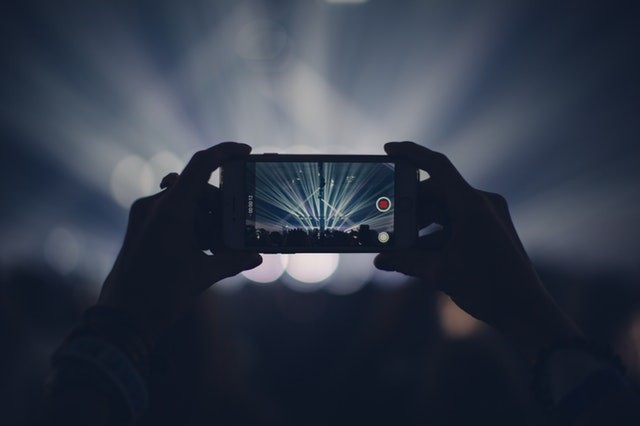 Person taking a photo of Play City landmark at night with flash photography, featuring a video game console and a mobile phone under a cloudy sky.
