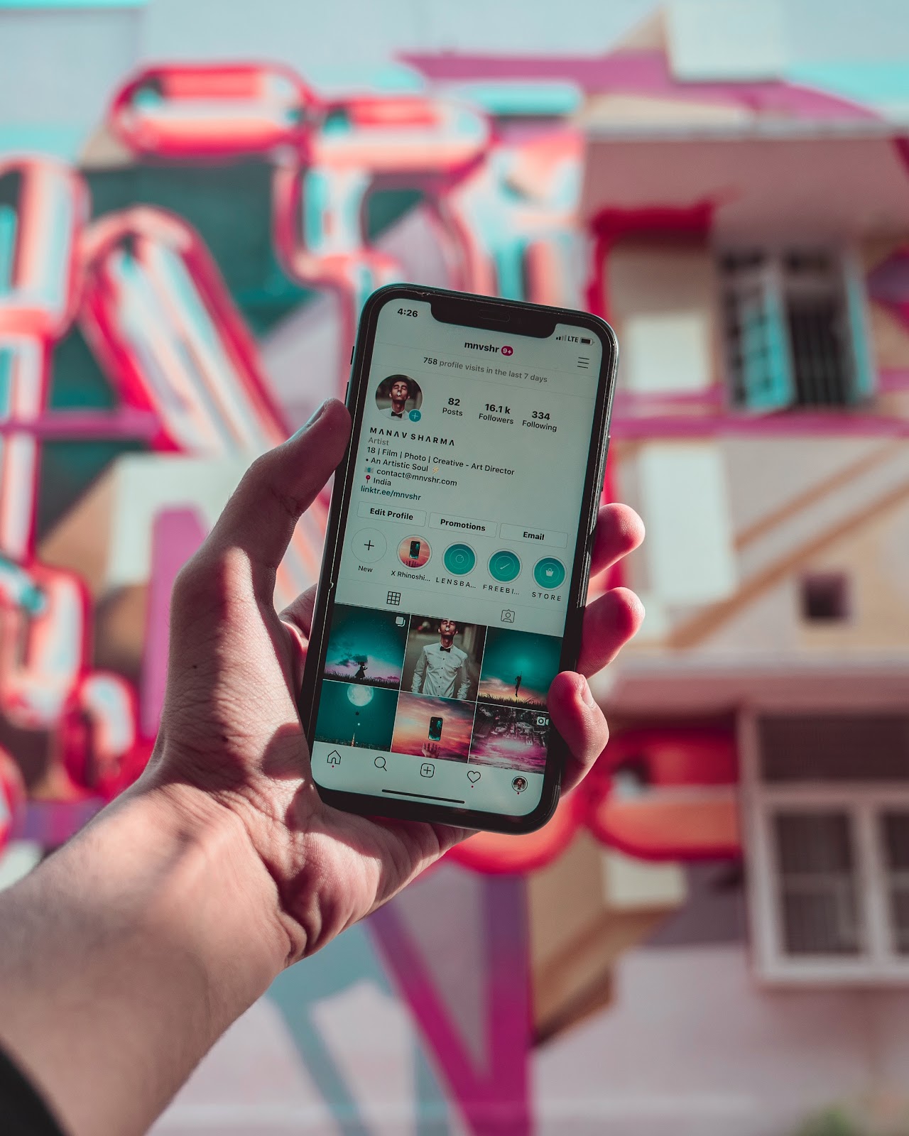 Hand holding a mobile phone displaying text: "4:26 mnvshr 9+ 758 profile visits in the last 7 days LTE 82 Posts: 16.1k Followers Following 334 MANAV SHARMA Artist 18