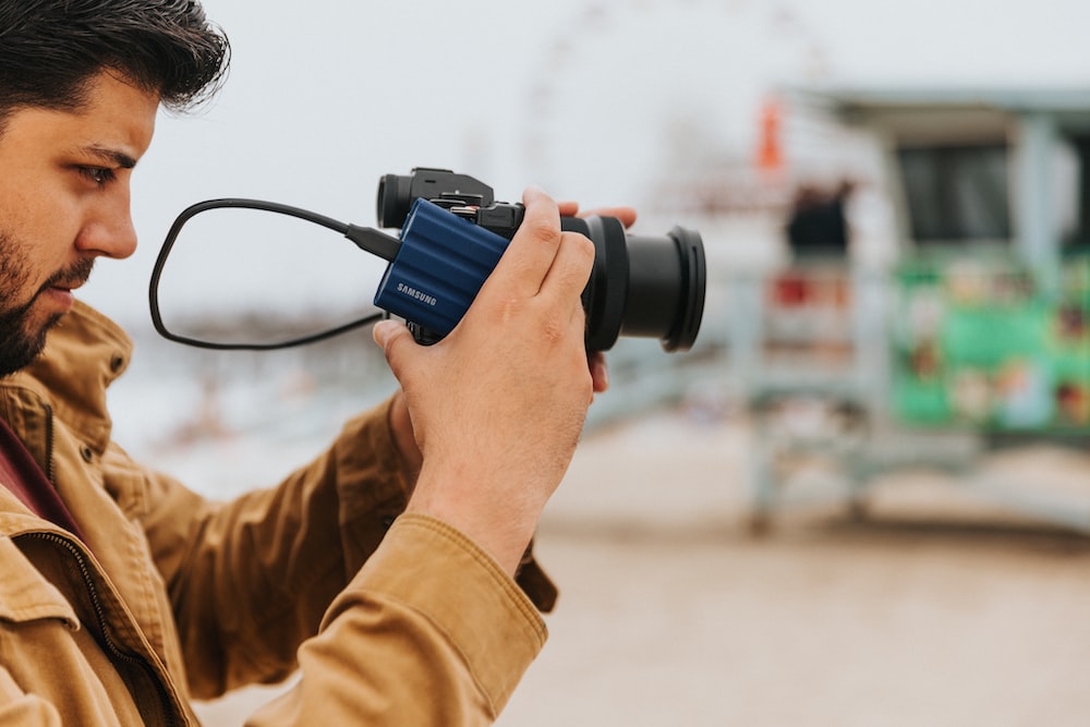 A person holding a Samsung digital camera with camera lens, wearing a jacket, in front of a single-lens reflex camera.