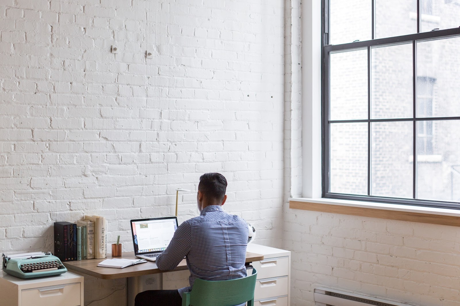 Person sitting at a computer desk near a window overlooking a building, wearing a shirt, with a laptop on the desk.