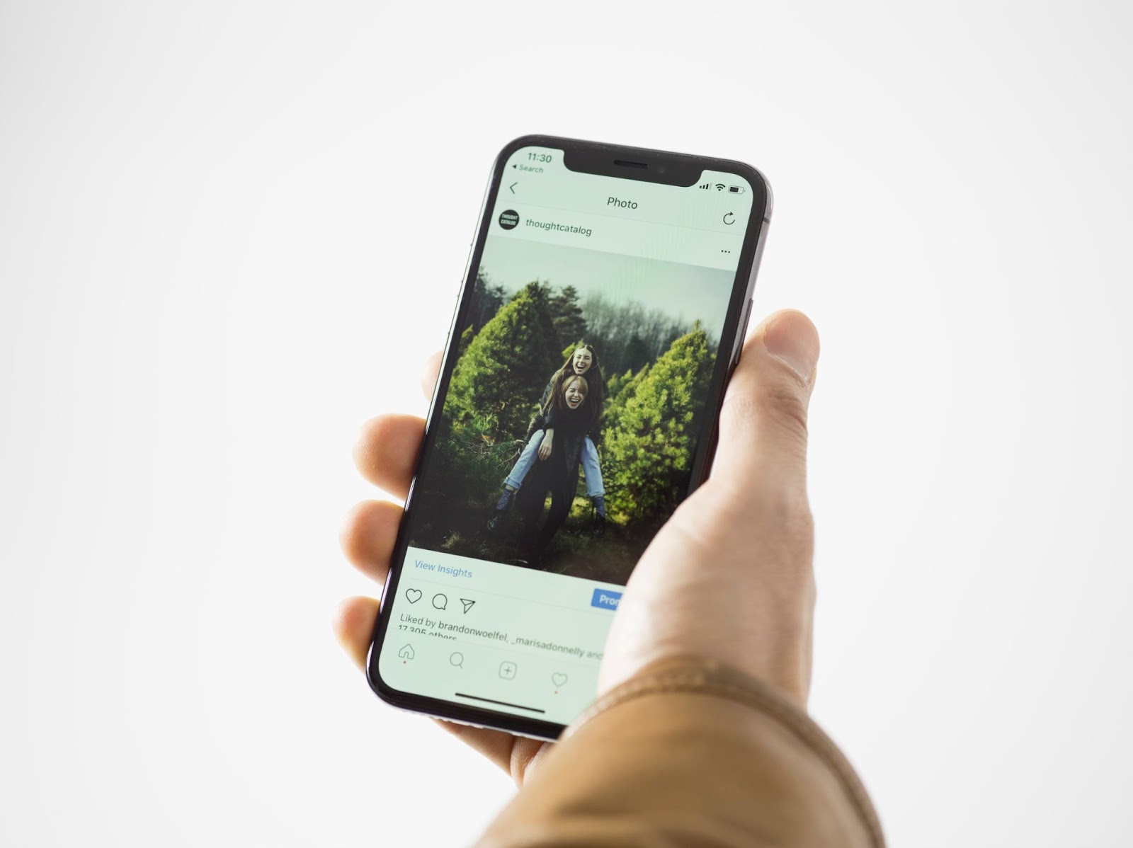 Hand holding a mobile phone displaying a social media analytics dashboard with the time 11:30, search bar, thoughtcatalog text, photo icon, insights, and likes from brandonwoelfel and others.