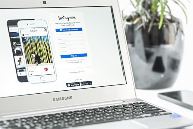 Computer, laptop, and mobile phone next to a houseplant, displaying Instagram logo and text to sign up on the App Store.