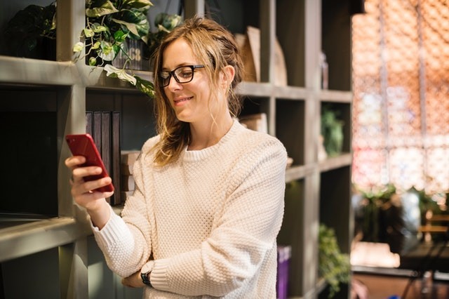 A person wearing glasses and smiling, holding a mobile phone, with a plant in the background.